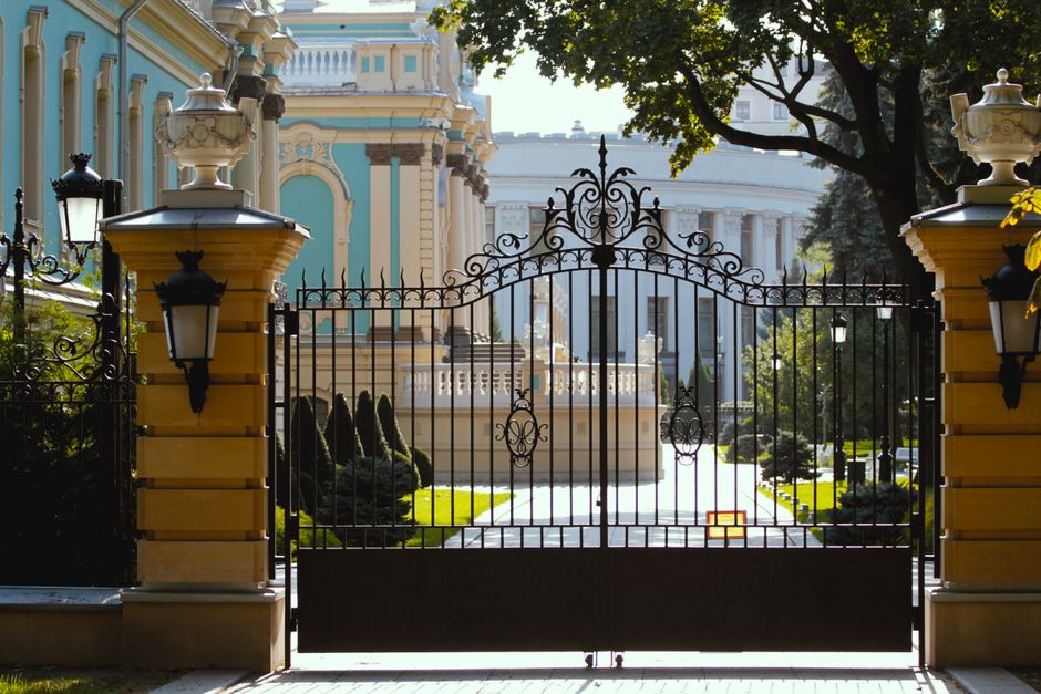 Ornate iron gate leading to historic architecture in a sunlit, lush setting.